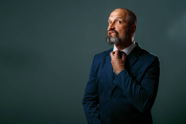 Bald middle-aged man with a beard, in a jacket with a white shirt straightens his blue tie on a dirty gray background - Foto, immagini