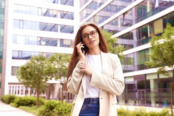 Portrait of young businesswoman talking with somebody on her mobile phone while walking on the street between office buildings.  - Foto, Imagem
