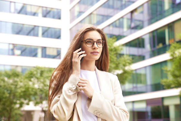 Portrait of young businesswoman talking with somebody on her mobile phone while walking on the street between office buildings.  - Photo, Image