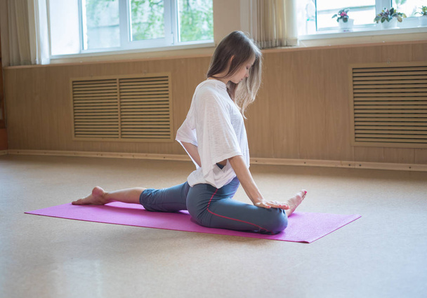 Young slim woman with blonde hair sitting on the yoga mat and doing stretching exercises - Photo, Image