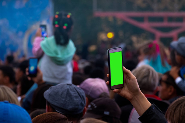 Eine weibliche Hand mit einem Smartphone nimmt auf oder überträgt ein Live-Konzert inmitten einer Menge von Fans. ein Leerzeichen zum Einbetten eines beliebigen Bildes anstelle eines grünen Bildschirms. Video auf dem Handy als Vorlage, Attrappe. - Foto, Bild