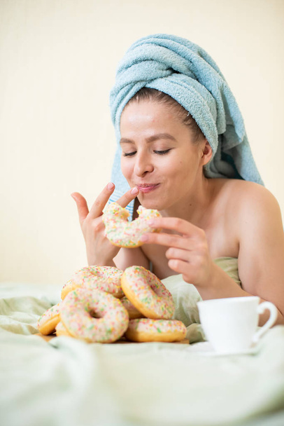 The girl in a towel has breakfast in the bed. A young attractive woman eats donuts in bed. The girl with a towel on her head happily eats donuts. - Fotoğraf, Görsel