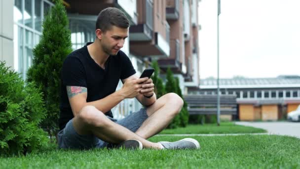 魅力的な男性は、ホテルの近くの草の上に座って、友人とおしゃべりしながら、携帯電話を使用します - 映像、動画