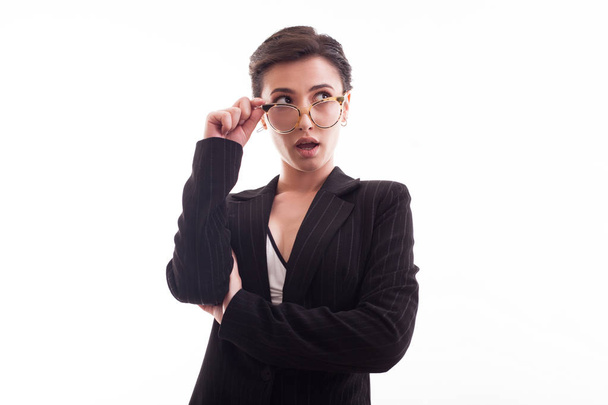 Stylish young fashion model holding her glasses and wearing a black jacket over white background in studio - Foto, afbeelding