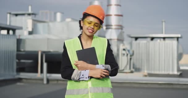 На даху будівельного майданчика посміхаючись велика Африканська жінка молодого інженера, що дивиться прямо на камеру носіння жовтого захисту окуляри і помаранчевий шолом - Кадри, відео