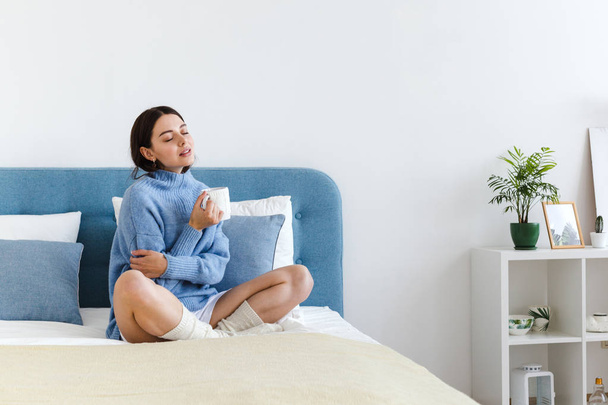 Girl in a blue sweater in interior Hygge style with a cup in hand sits on the bed - Foto, Imagem