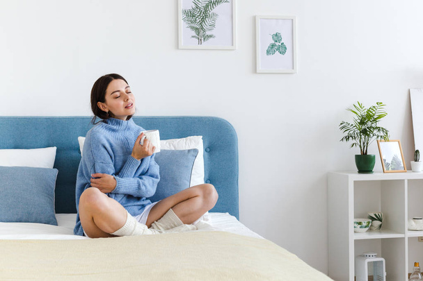 Girl in a blue sweater in interior Hygge style with a cup in hand sits on the bed - 写真・画像