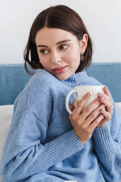 Beautiful face of a girl, a girl in blue sweater sits on a sofa with a cup of hot tea in her hands - Fotoğraf, Görsel