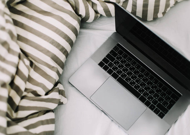 Laptop in bed on white linens. Work at home concept.  Flat lay, top view, lifestyle, minimalist workspace background.  - 写真・画像