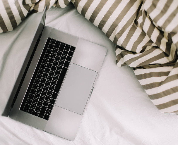Laptop in bed on white linens. Work at home concept.  Flat lay, top view, lifestyle, minimalist workspace background.  - Φωτογραφία, εικόνα