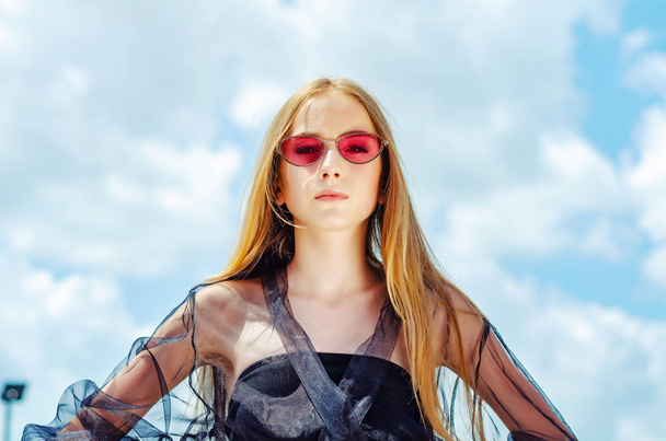 Chica joven en gafas de sol rojas posando sobre el fondo del cielo, brillante
  - Foto, Imagen