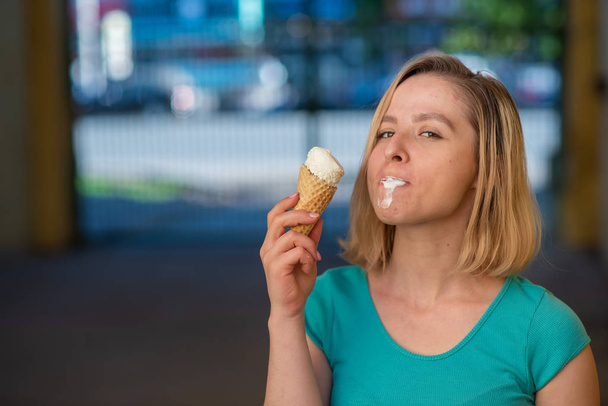 Portrait of a cute girl in a green dress is walking outside and eating dessert. Beautiful blonde enjoying a cone with ice cream on a beautiful summer day. Life is a pleasure - Zdjęcie, obraz