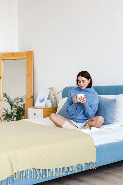 Lány egy kék pulóvert belső Hygge stílusban egy csésze kézzel ül az ágyon - Fotó, kép