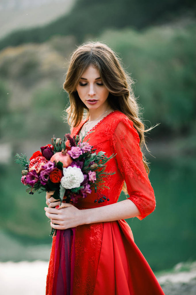 Young bride in a red dress with an Assemtric bouquet of pomegranates and roses. - Photo, Image