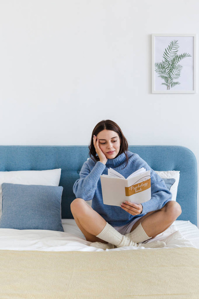 Girl in a blue sweater reading a book on psychology sitting on the bed in a cozy interior - Fotografie, Obrázek