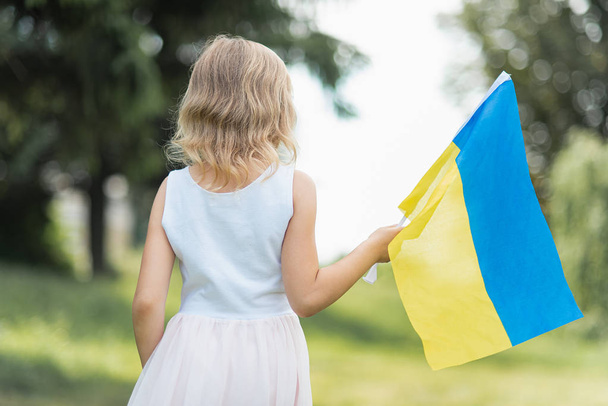 back view of little girl with fluttering blue and yellow flag of Ukraine walking in field at sunny summer day, Ukraine Independence Day concept - Foto, Imagem