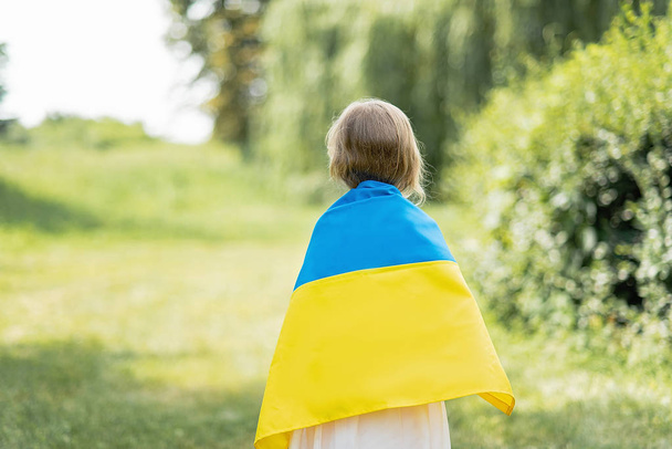 back view of little girl carrying fluttering blue and yellow flag of Ukraine on shoulders in field at sunny summer day, Ukraine Independence Day concept - Fotoğraf, Görsel