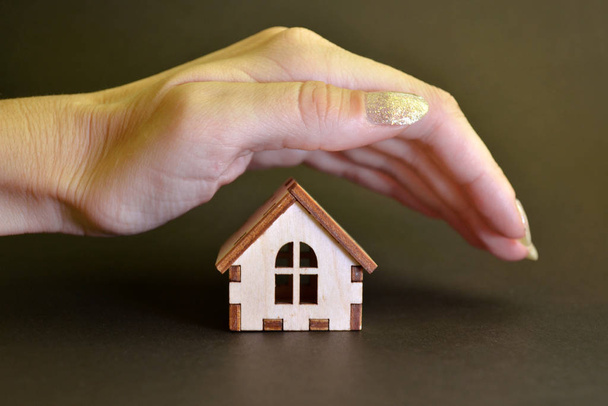 A wooden toy house stands on a black background and a female hand closes it from above, house protection concept - Photo, Image