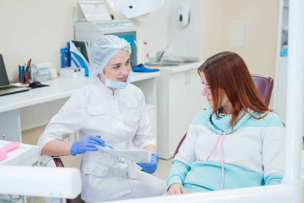 Young beautiful female dentist talking to a patient in the office. A red-haired woman sits in a chair with an orthodontist and listens to appointments. Dental services - 写真・画像