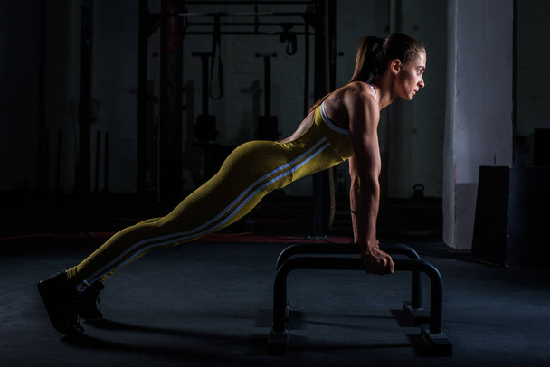 Young athletic girl in yellow tight sports uniform doing horizontal push-ups with bars in gym - Foto, Imagen