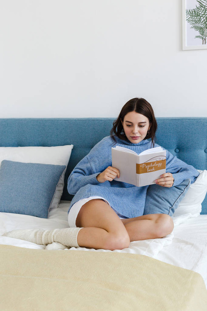 Girl in a blue sweater reading a book on psychology sitting on the bed in a cozy interior - Foto, immagini