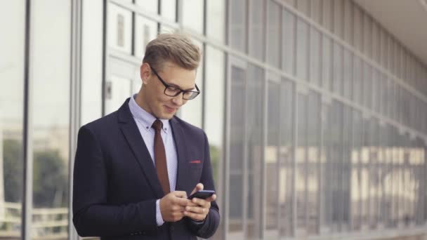 Happy businessman smiling as he reads a text message standing outside the office. Male executive manager in trendy clothes using smartphone outdoors. Modern technology concept - Metraje, vídeo