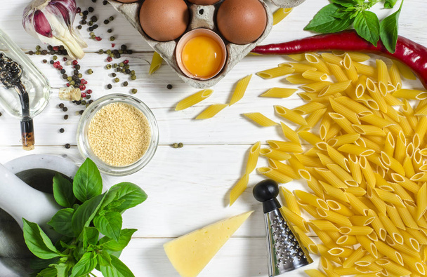 Penne pasta, groene basilicum, Parmezaanse kaas, sesam, knoflook, Chili, eieren liggen op een witte boom tafel - Foto, afbeelding