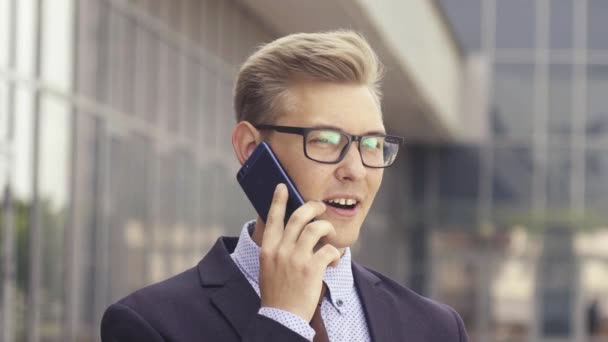 Jonge knappe Kaukasische zakenman in glazen praten op mobiele telefoon voor kantoorgebouw. Mannelijke Executive Manager in trendy kleding met behulp van smartphone buitenshuis. Modern technologie concept - Video