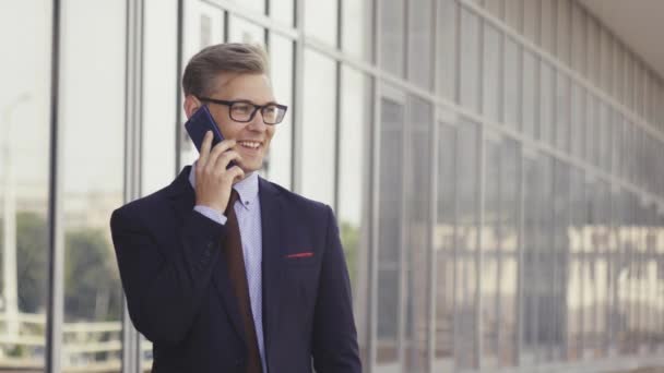Young handsome Caucasian businessman in glasses talking on mobile phone in front of office building. Male executive manager in trendy clothes using smartphone outdoors. Modern technology concept - Video