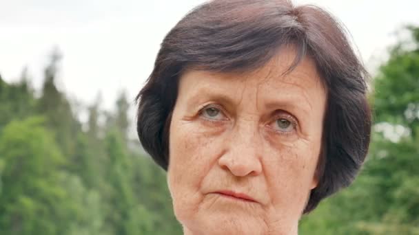 Portrait of thoughtful sad senior woman with short dark hair and wrinkles on her face on mountain hill with green forest on background - Footage, Video