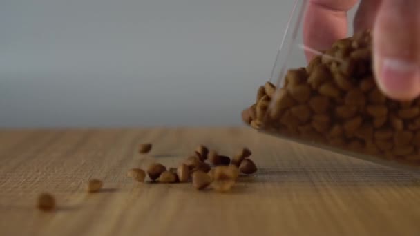 A mans hand sprinkles crispy dry cat food on a wooden surface from a glass cup - Footage, Video
