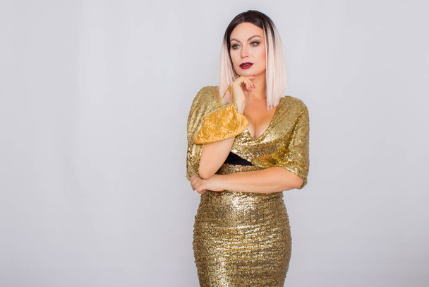 Charming young blonde haired woman wearing elegant gold evening dress and holding gold color clutch in her hands - 写真・画像