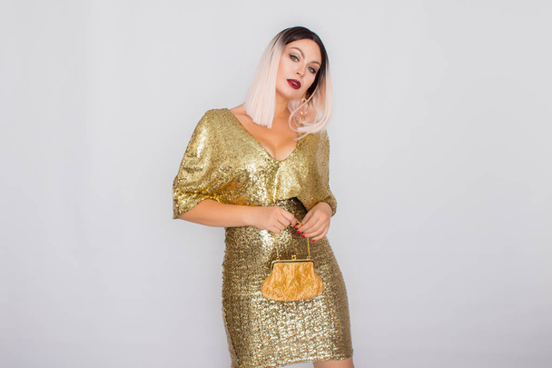 Charming young blonde haired woman wearing elegant gold evening dress and holding gold color clutch in her hands - Fotografie, Obrázek