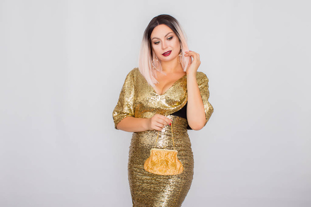 Charming young blonde haired woman wearing elegant gold evening dress and holding gold color clutch in her hands - Foto, afbeelding