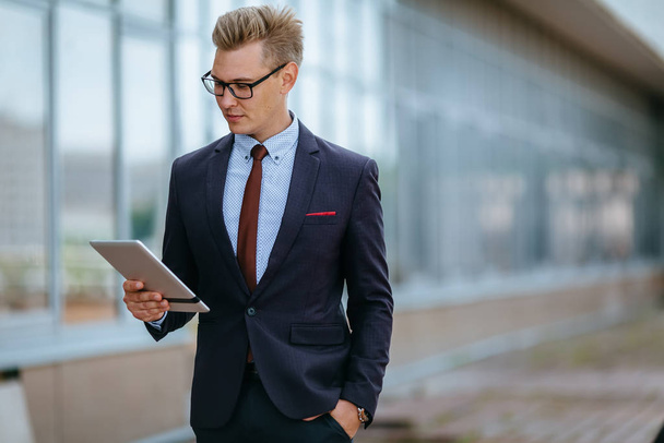 Elegant young businessman using his PC tablet. Portrait of a Male executive manager in trendy clothes with tablet pc in front of office building. Modern technology concept - 写真・画像