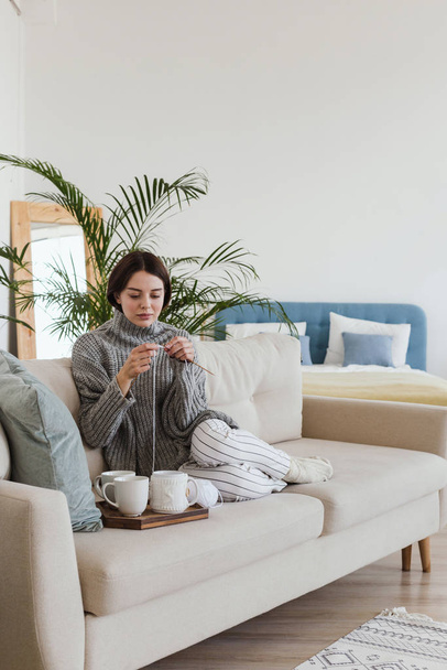 дівчина в теплому сірому светрі в'яжеться, сидячи на дивані в затишній внутрішній гігієні
 - Фото, зображення