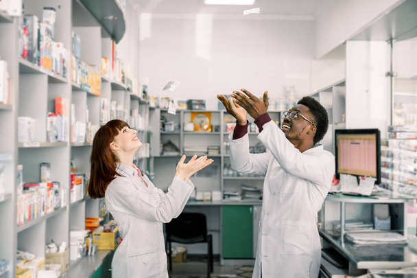 Two funny smiling young pharmacists having fun in pharmacy, throw up different pills - Foto, Imagen