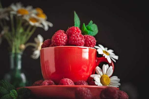 Raspberry in a red cup with chamomile and leaves on a dark background. Summer and healthy food concept. Selective focus. - Фото, зображення