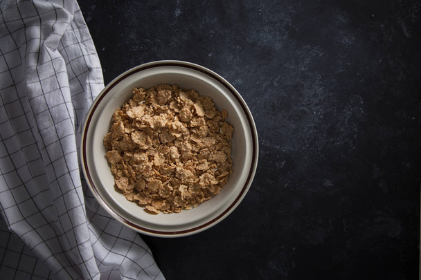 Salvado de trigo cereal de desayuno sin leche en un tazón. Fondo negro con servilleta hecha en casa
.  - Foto, imagen