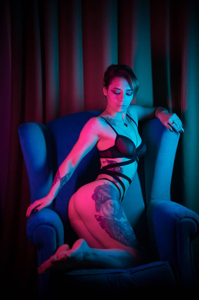 Молода жінка з багатьма татуюваннями в сексуальній нижній білизні, сидячи в кріслі в неоновому освітленні
 - Фото, зображення