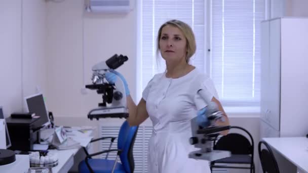 Beautiful blonde in medical gown dances in laboratory with microscopes - Footage, Video