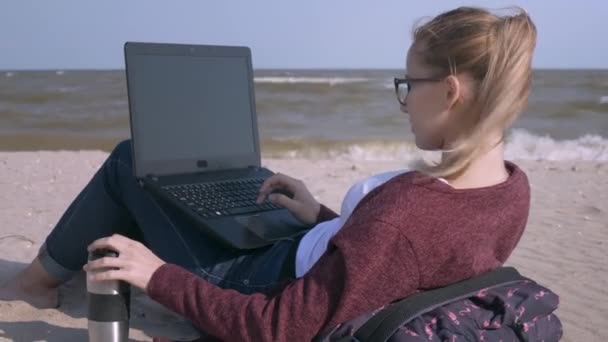 A fiatal, jóképű nő dolgozik a laptop és a kávéfogyasztás a strandon a beállítás nap. Fiatal üzletember dolgozik a tengerparton naplementekor. Szabadúszóként működik a tengerparton. - Felvétel, videó
