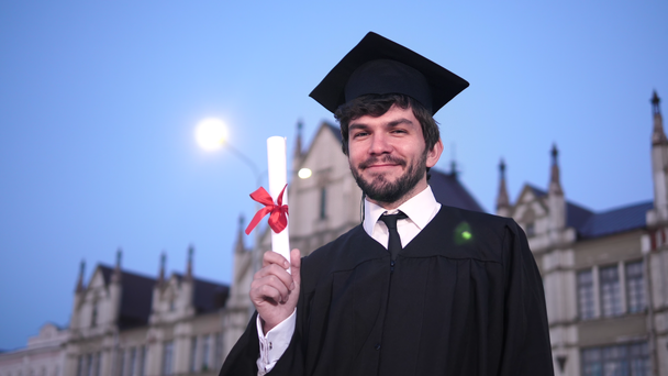 Guy a diplomaszerzés ruhát mosolyogva és mutatja diploma kamera. - Felvétel, videó
