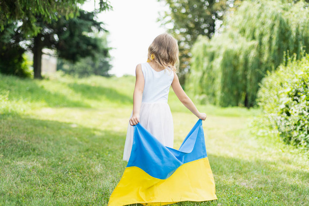 back view of little girl with fluttering blue and yellow flag of Ukraine walking in field at sunny summer day, Ukraine Independence Day concept - Fotografie, Obrázek