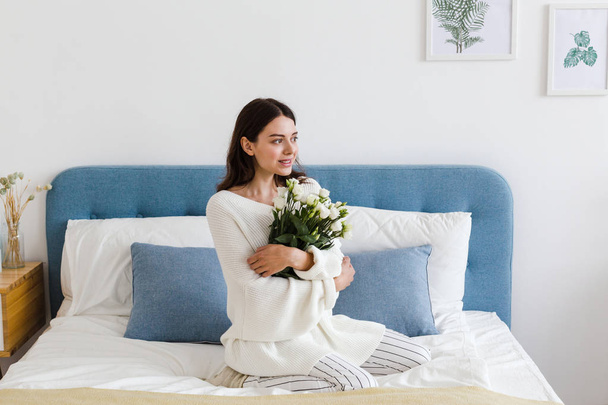 A girl in a white sweater sits on a bed holding a bouquet of white roses in her hand - Foto, Bild