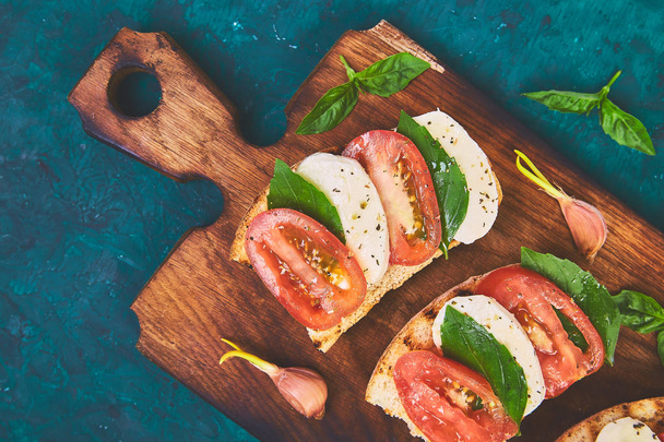 Caprese bruschetta toasts on cutting board. Bruschetta with tomatoes, mozzarella cheese and basil on a green background. Traditional italian appetizer or snack, antipasto - Photo, Image