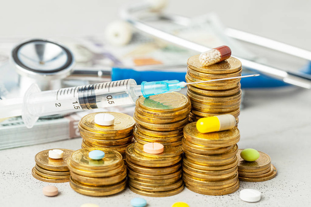 Stapel munten met pillen en spuit op de achtergrond van stethoscoop en cash geld. - Foto, afbeelding