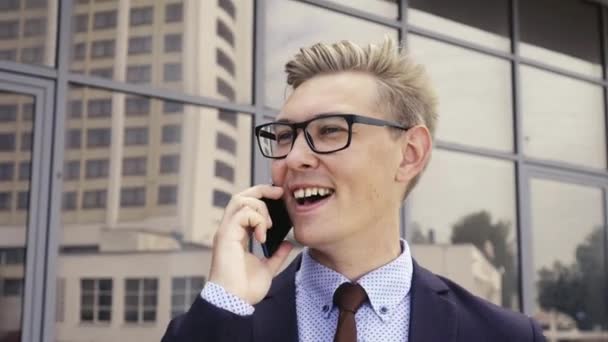 Young handsome Caucasian businessman in glasses talking on mobile phone in front of office building. Male executive manager in trendy clothes using smartphone outdoors. Modern technology concept - 映像、動画