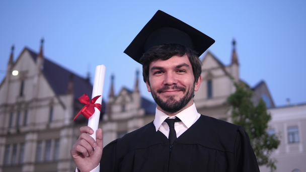 Kaukázusi boldog fiatal diplomás férfi pózol a kamerába, és bemutatja a diplomáját az egyetem előtt. - Felvétel, videó