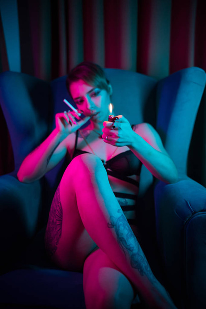 Молодая женщина в сексуальном белье сидит в кресле со скрещенными ногами и держит зажигалку - вот-вот зажжет сигарету
 - Фото, изображение